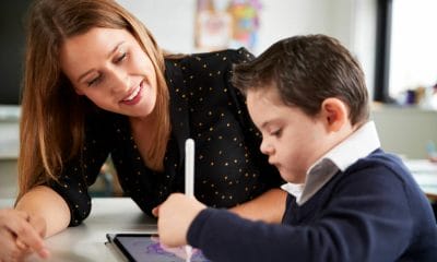 Cierre de una profesora sentada en el escritorio con un escolar con síndrome de Down usando una tablet computadora en un aula de la escuela primaria