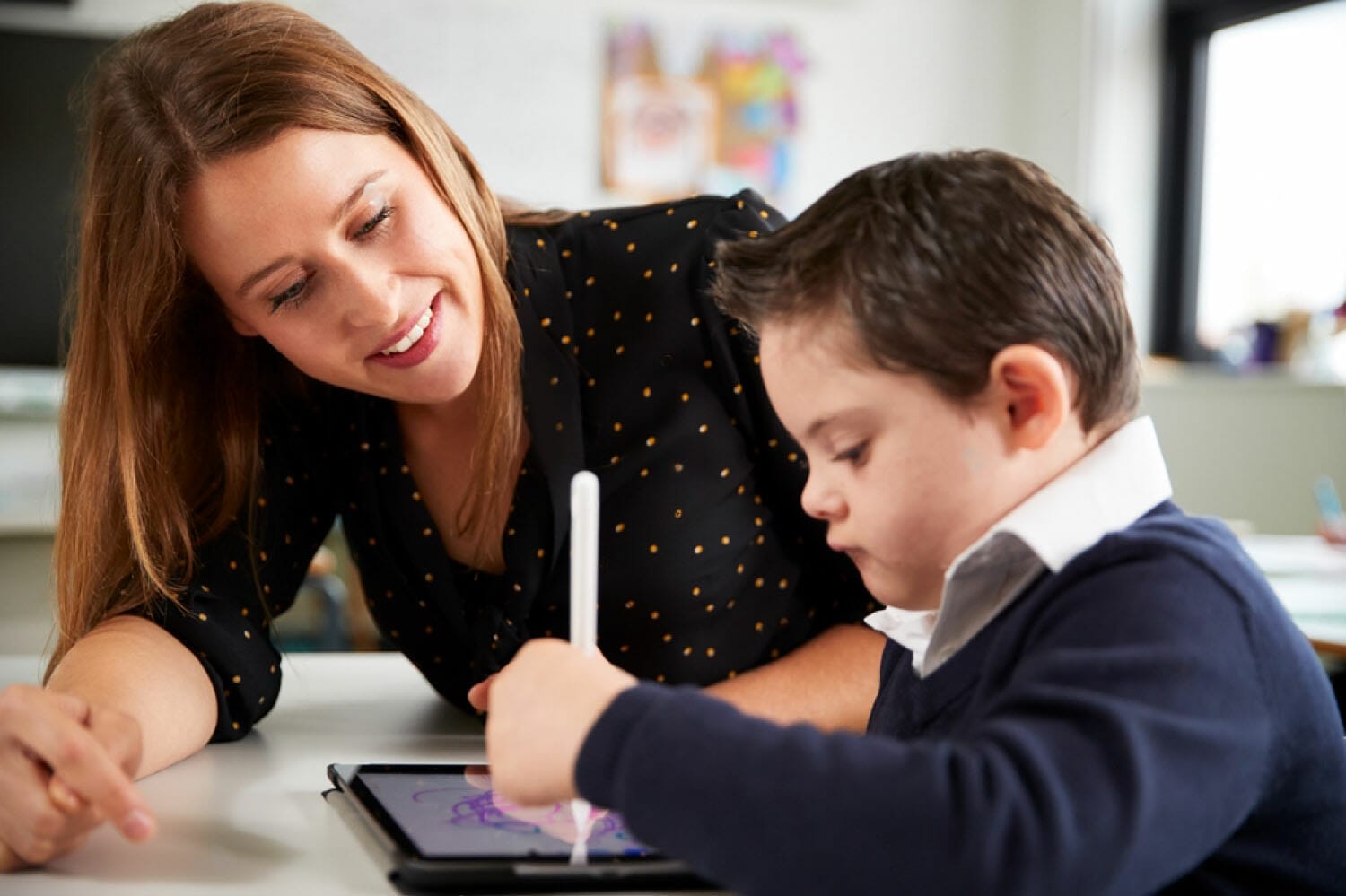 Cierre de una profesora sentada en el escritorio con un escolar con síndrome de Down usando una tablet computadora en un aula de la escuela primaria